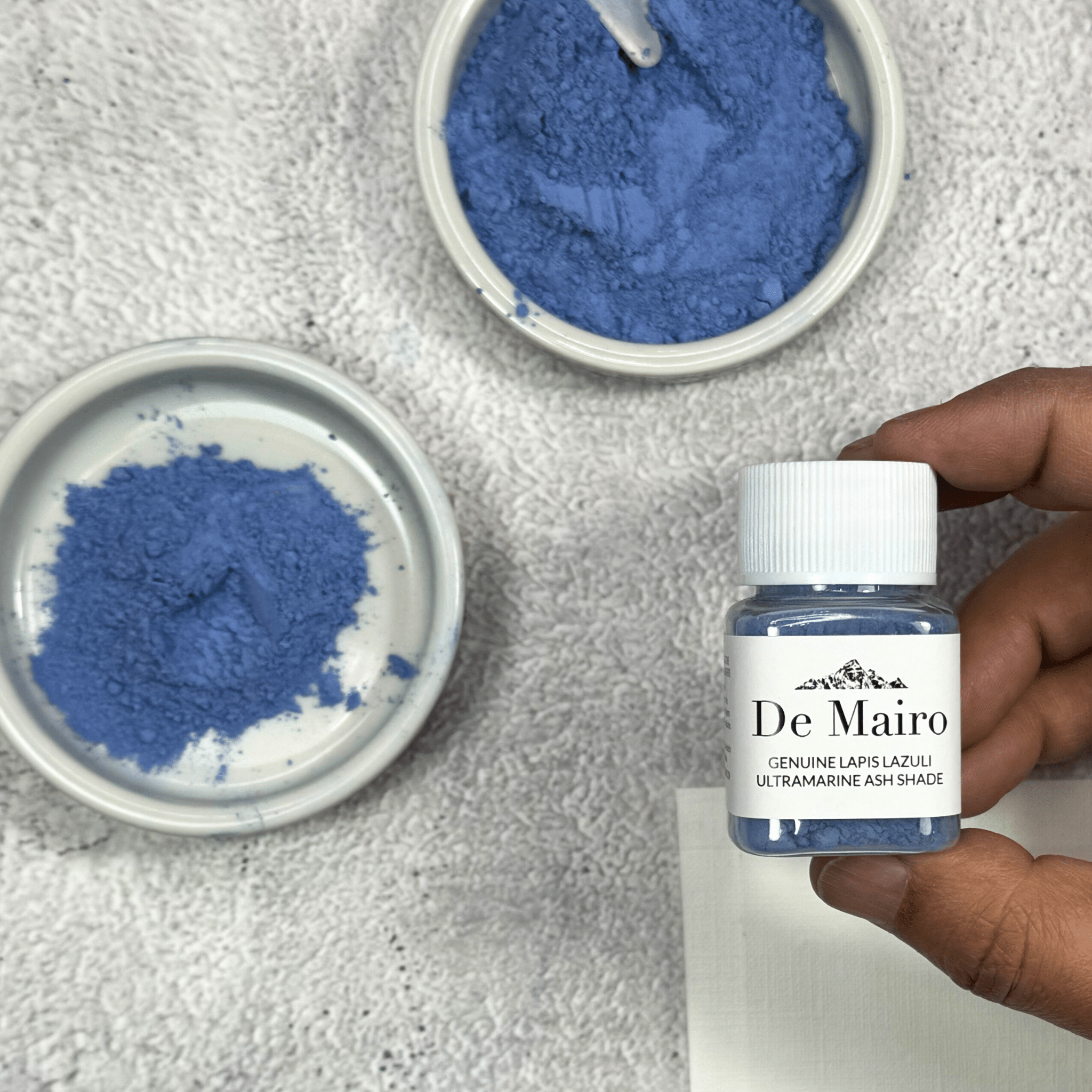Genuine Lapis Lazuli Ultramarine Ash Shade Pigment jar holding in fingers over dishes of dry pigment