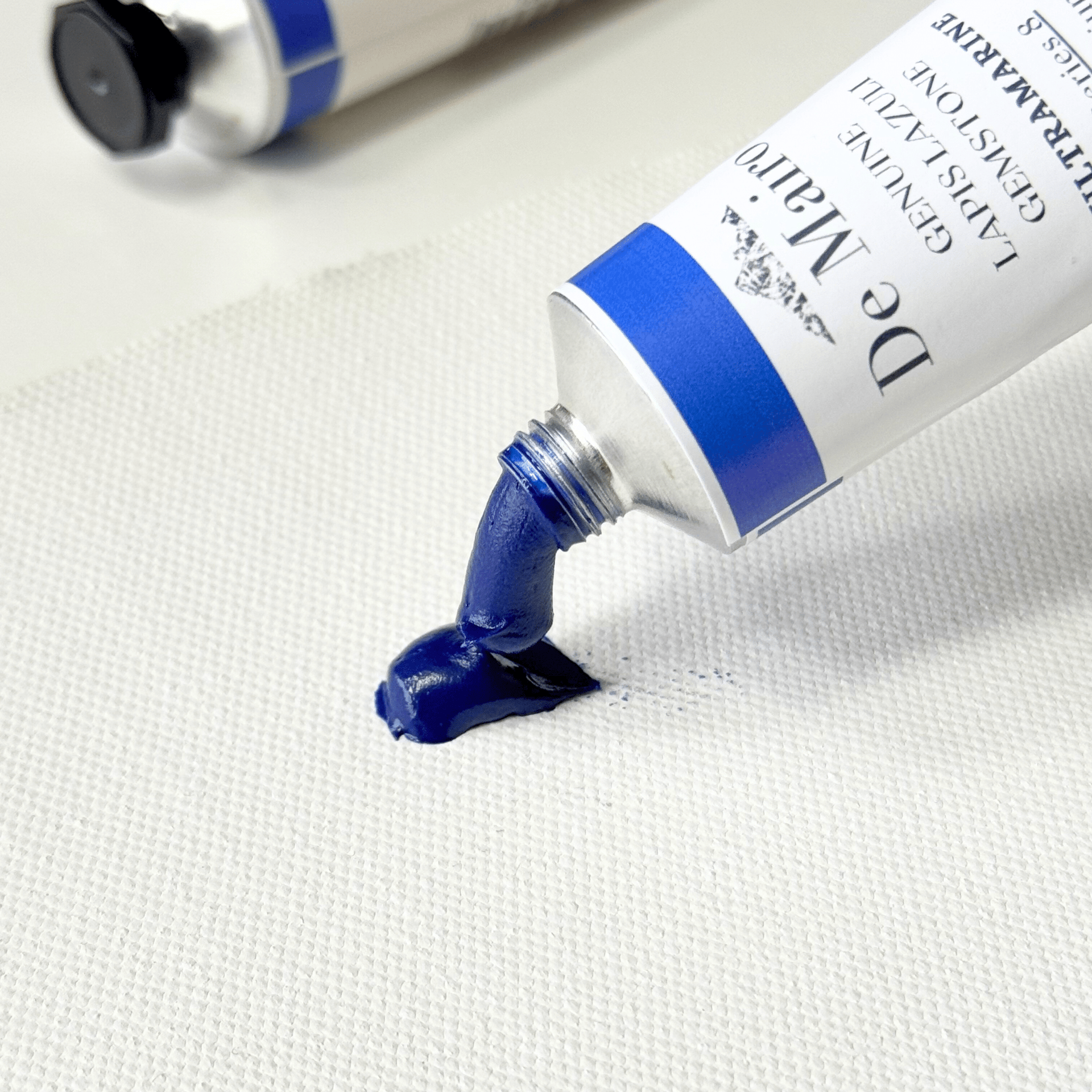 A close-up of a tube of De Mairo Ultramarine blue oil paint being squeezed onto a white textured canvas