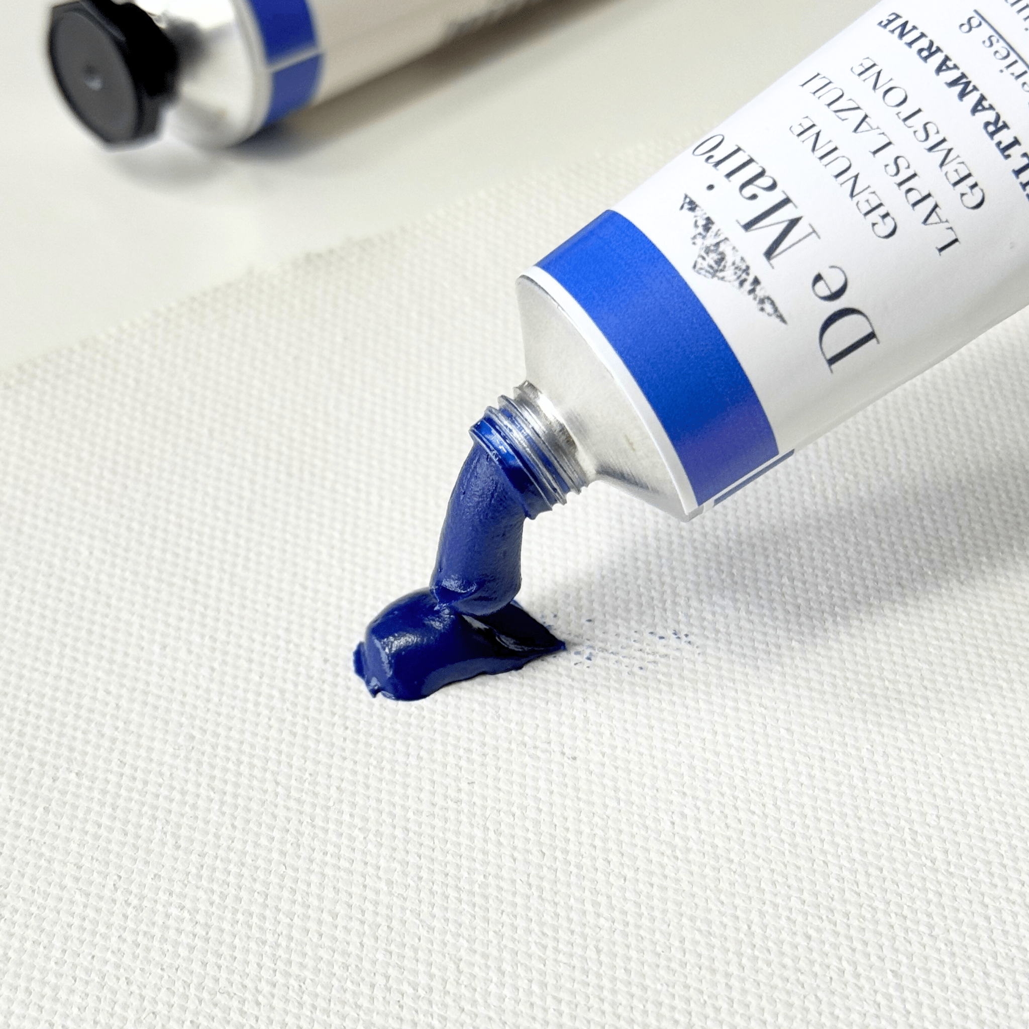 A close-up of a tube of De Mairo Ultramarine blue oil paint being squeezed onto a white textured canvas