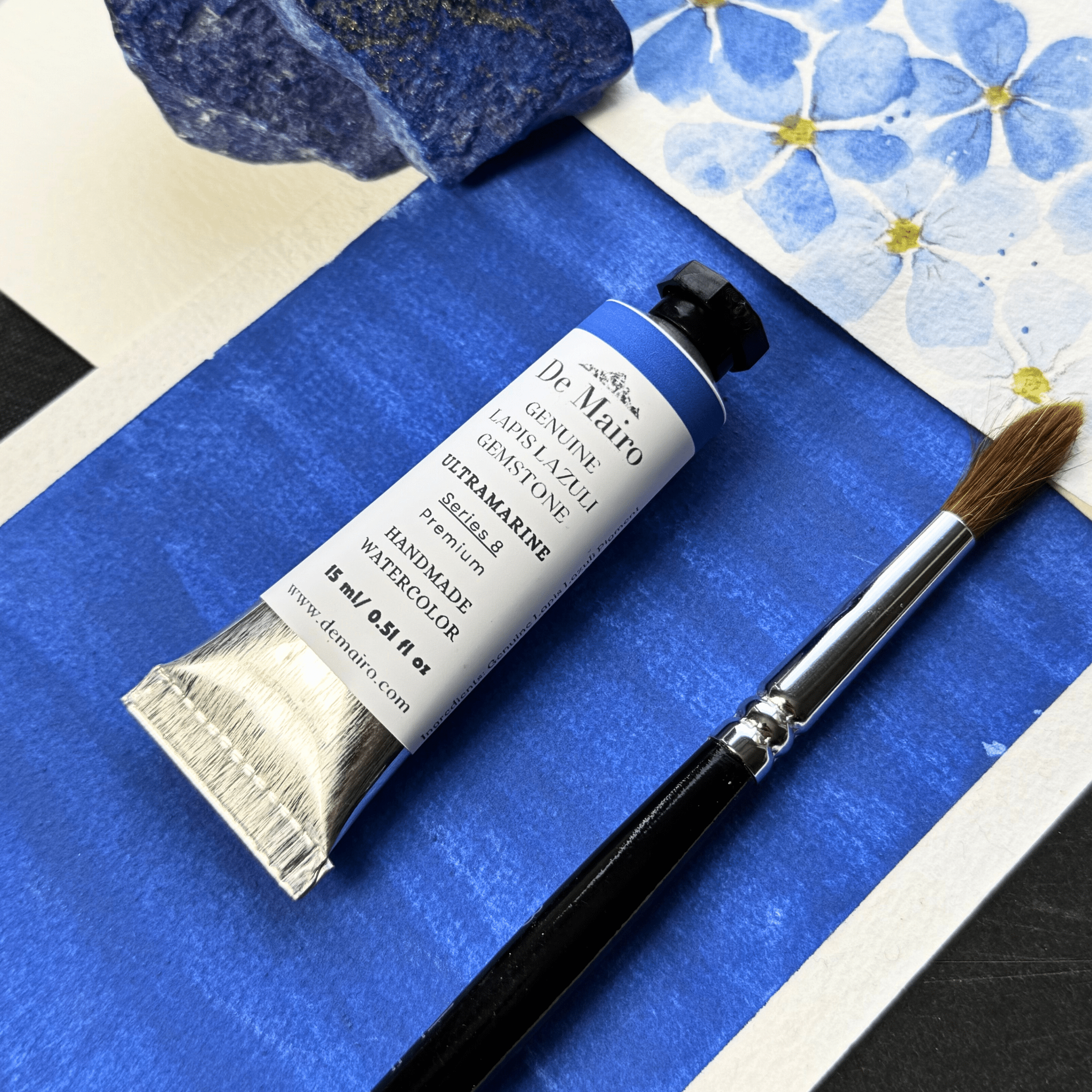Tube of De Miero watercolor paint and a brush on a blue surface with floral pattern alongside a gemstone