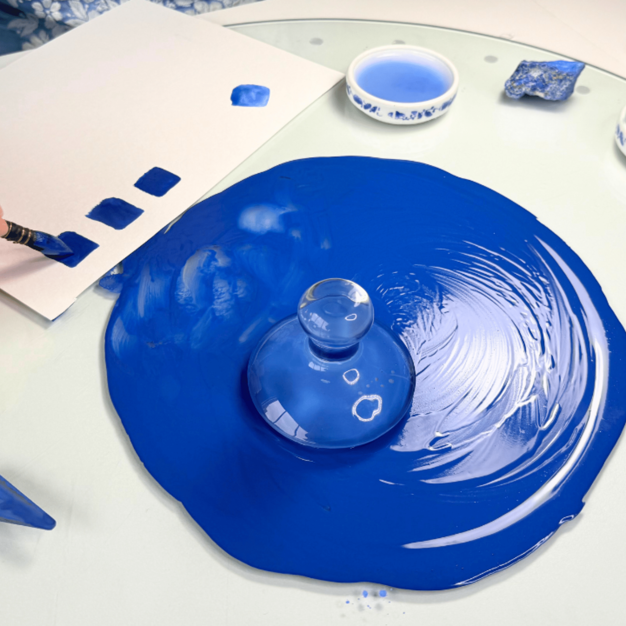 Blue glass lid on a white surface with a brush and blue paint.