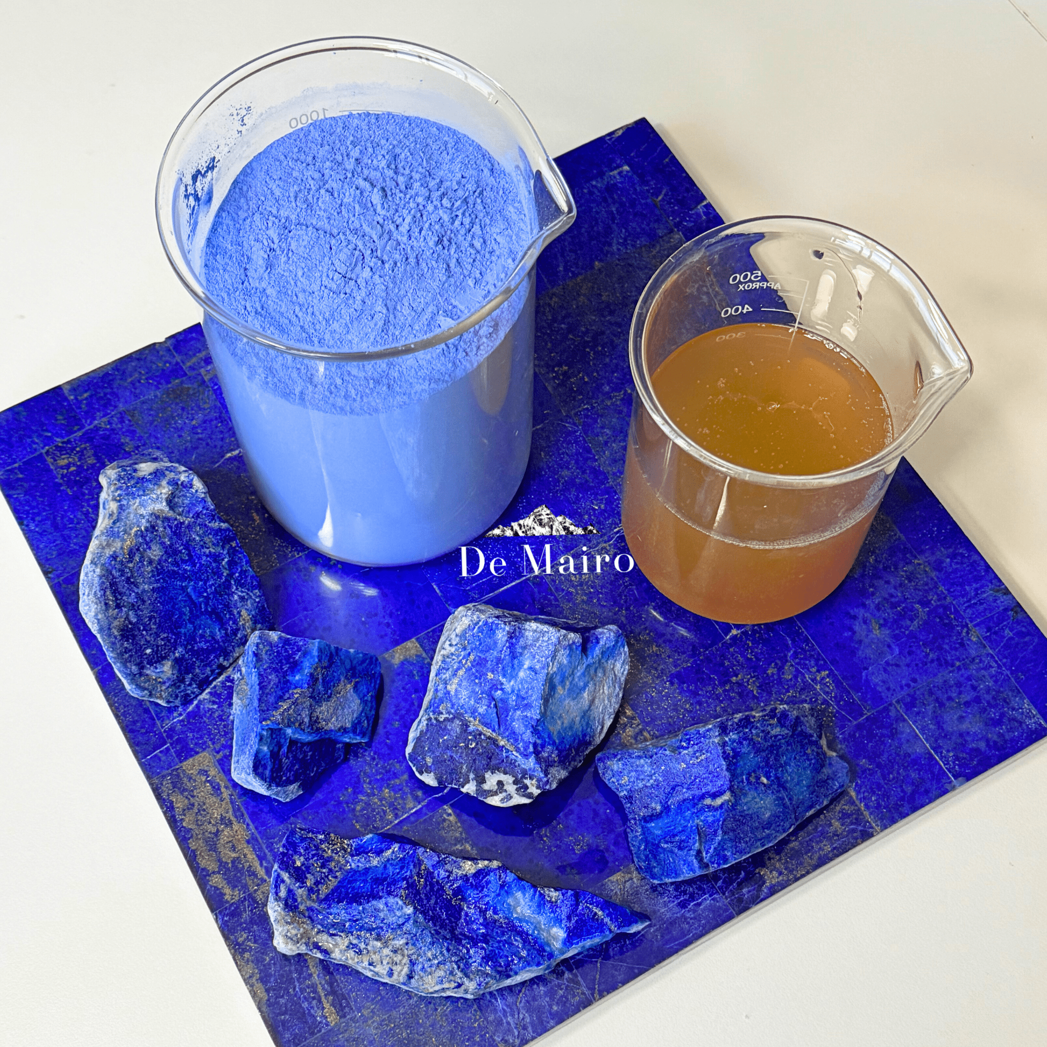 Two beakers with blue powder and brown liquid on a blue surface beside lapis lazuli stones with 'De Mairo' branding.