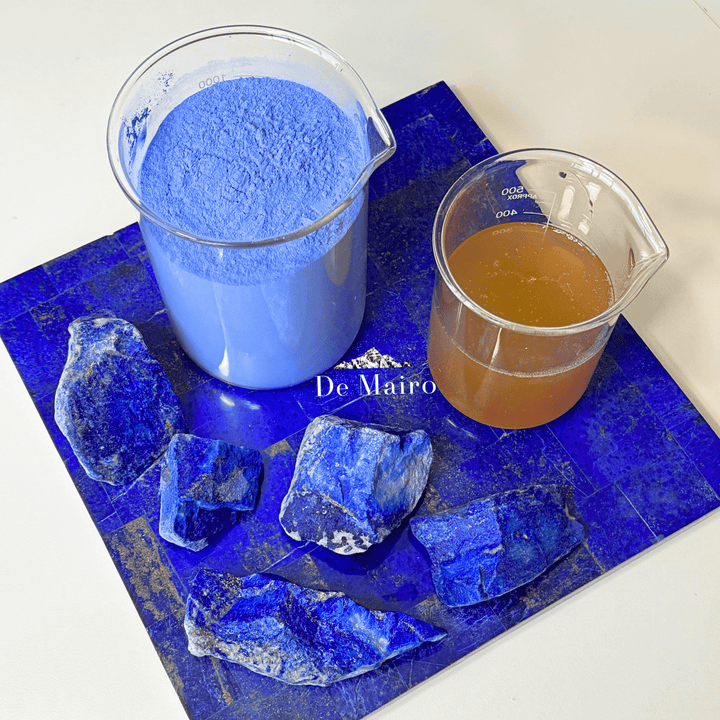 Two beakers with blue powder and brown liquid on a blue surface beside lapis lazuli stones with 'De Mairo' branding.
