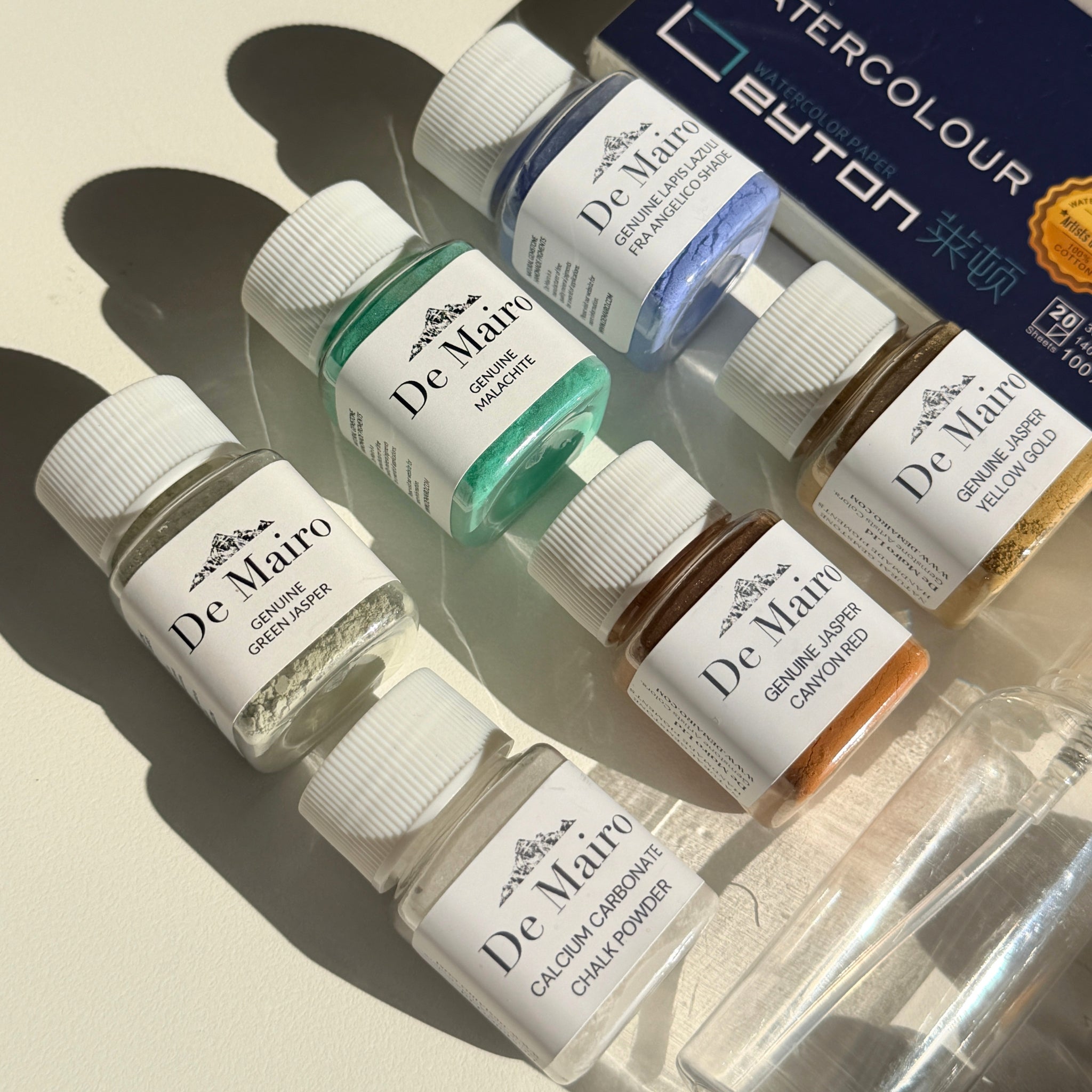 De Mairo genuine gemstone pigment and medium bottles in natural light with watercolour block