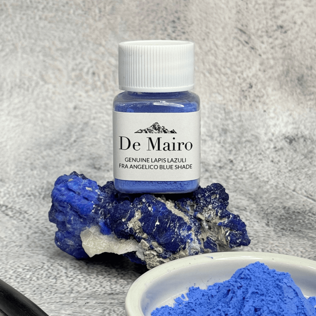 Jar of De Mairo genuine lapis lazuli Fra Angelico blue pigment resting on a raw lapis stone beside a dish of blue powder