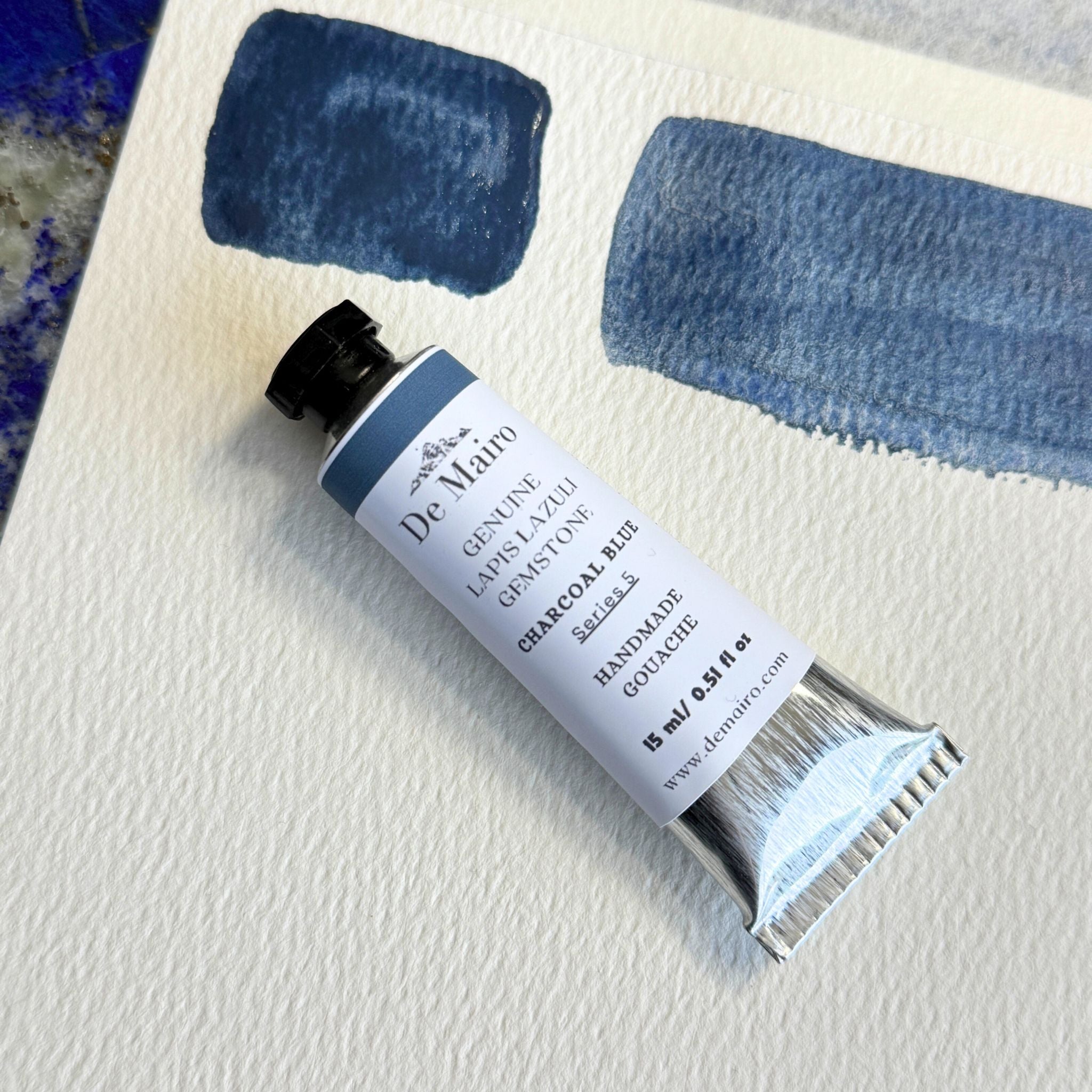Tube of De Maira charcoal pastel on a textured surface with blue color swatches.