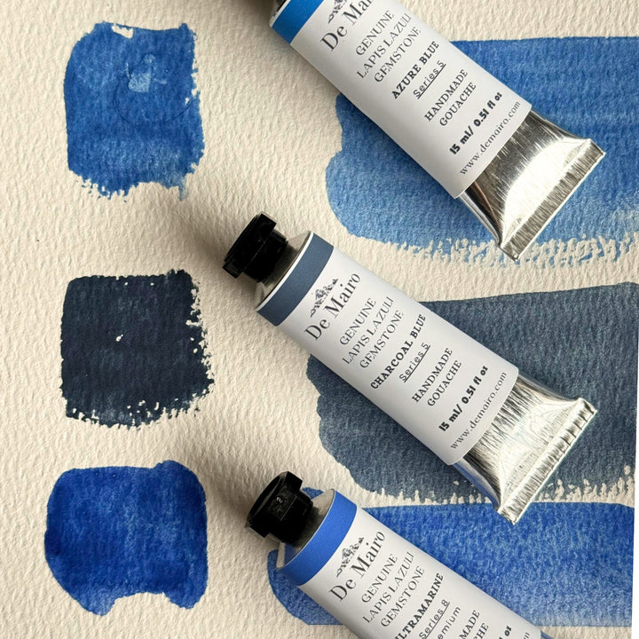 Three tubes of De Mairo blue gouache shades on a textured white surface with blue paint swatches.