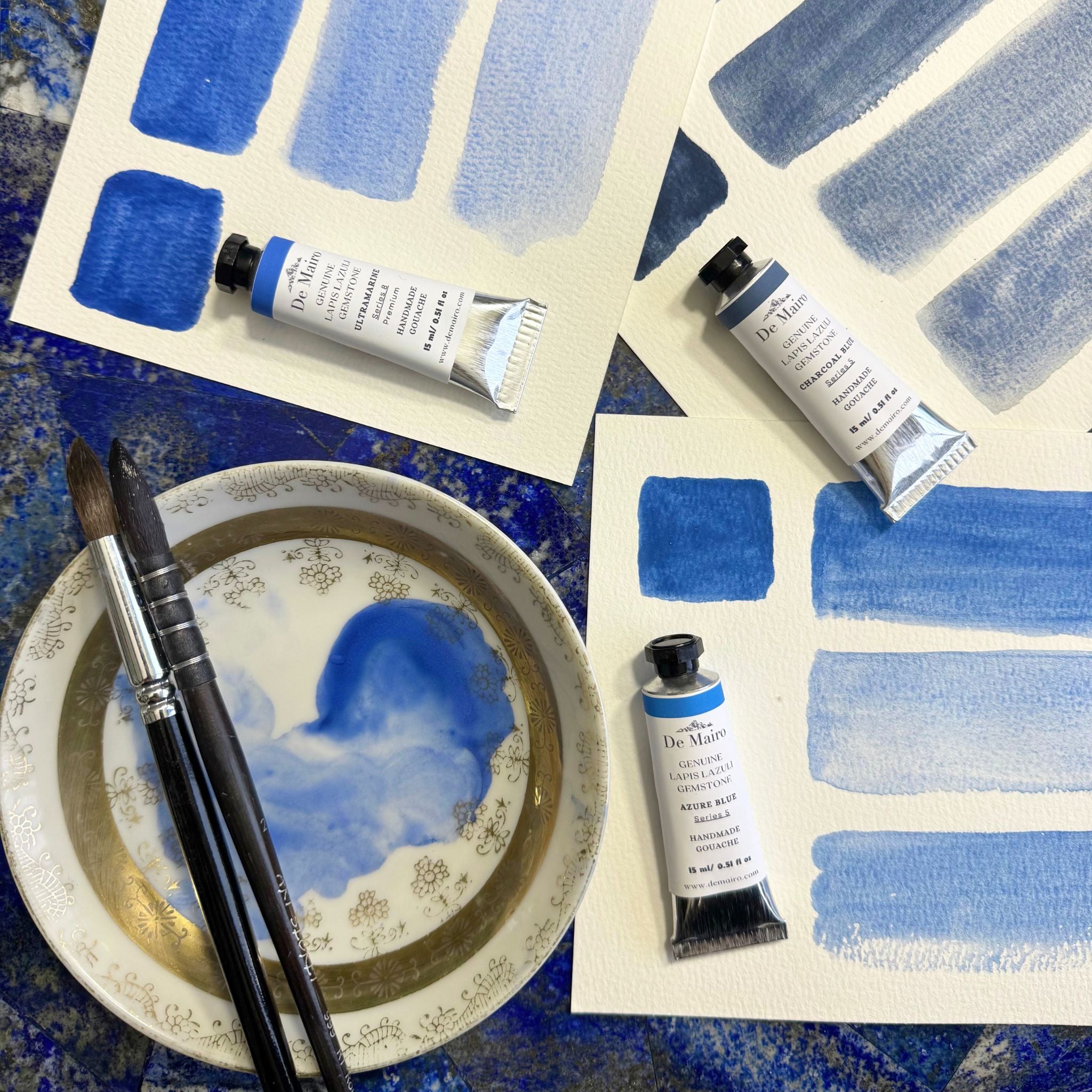 De mairo gouache tubes with blue brush strokes on paper and a bowl with paint.