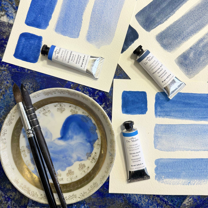 De mairo gouache tubes with blue brush strokes on paper and a bowl with paint.